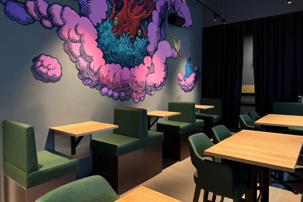 Sitteområde med grønne stoler og trebord foran en fargerik veggmaleri hos Annis Pølsemakeri.