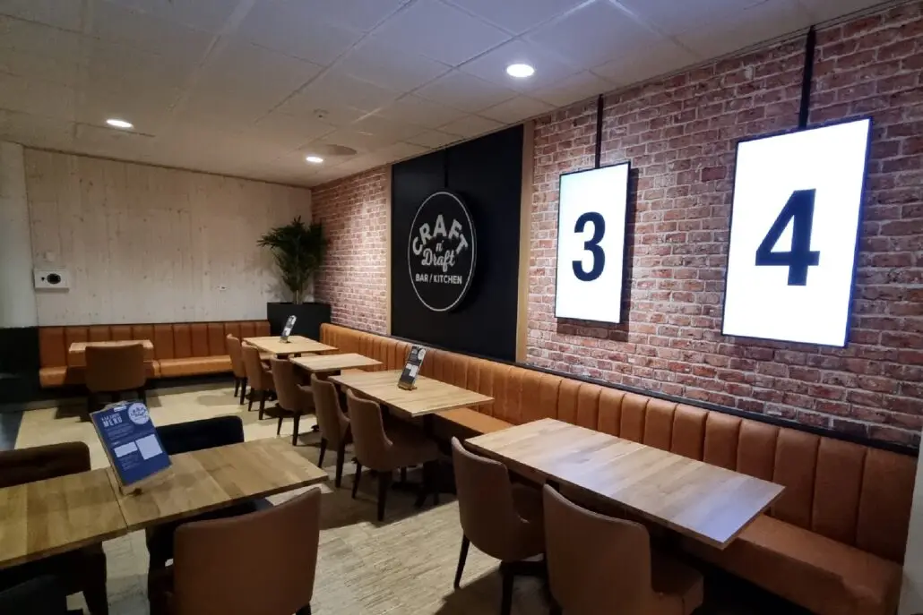 Sitteområde hos Craft’n Draft med mursteinsvegg, skilt med logo, store tallskilt og trebord med skinnbenker.