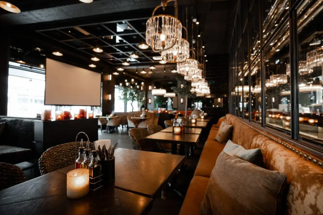 Restaurantinteriør på Olivia Lillestrøm med myke sofaer, mørke trebord, elegante lysekroner og en stor skjerm til arrangementer.