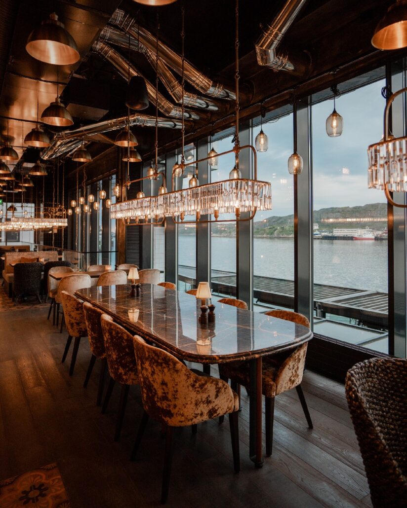 Elegant spiseområde på Olivia Ramsalt med store vinduer mot sjøen, lange marmorbord, fløyelsstoler og moderne krystall-lamper hengende fra taket.