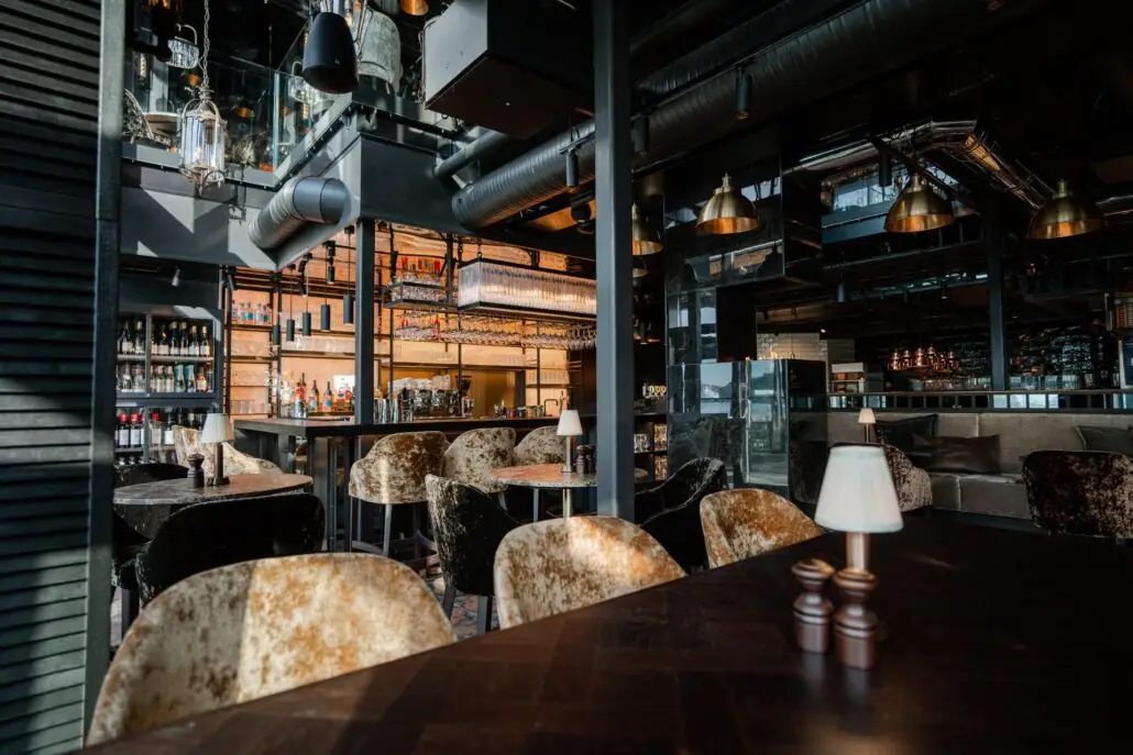 Interiør med barområde, fløyelsstoler og bordlamper. Hyller med vin- og brennevinsflasker i bakgrunnen og en moderne bar med hengende glass og flasker.