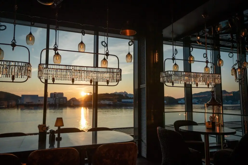 Restaurantbord ved store vinduer med utsikt mot sjøen og en solnedgang. Glasslamper henger fra taket og speiler det varme lyset fra solen som går ned bak horisonten.