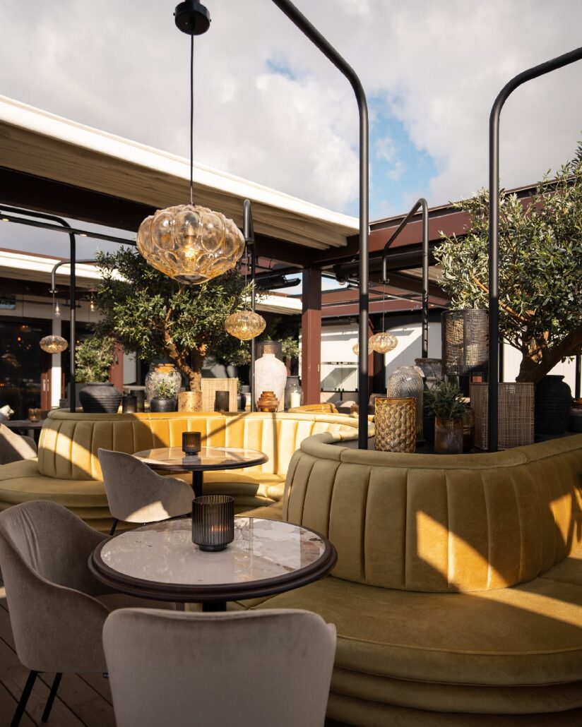 Uteservering på Olivia Vågen med gule buede sofaer, små runde marmorbord og beige stoler. Grønne oliventrær, dekorative lamper og keramikk skaper en stemningsfull og middelhavsinspirert atmosfære under åpen himmel.